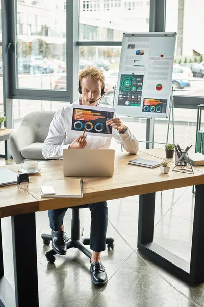 A redheaded professional in a white shirt engages in a project discussion using his laptop and headset. — Stock Photo