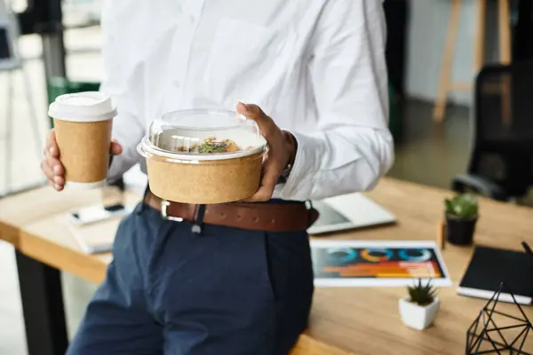 Ein Geschäftsmann balanciert eine Kaffeetasse und einen Lunchcontainer, während er sich in einem eleganten Büro auf sein Projekt konzentriert. — Stockfoto