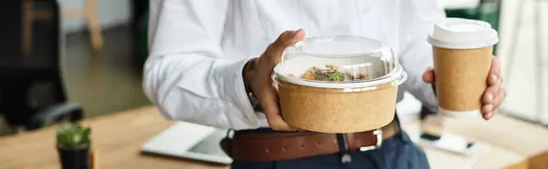 A businessman in a white shirt is engaged in work, balancing lunch and coffee in a modern workspace. — Stock Photo