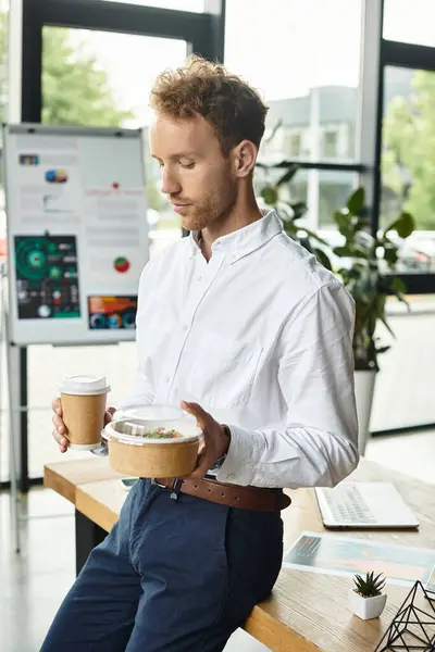A redhead businessman in a white shirt holding lunch and coffee in a modern office. — Stock Photo