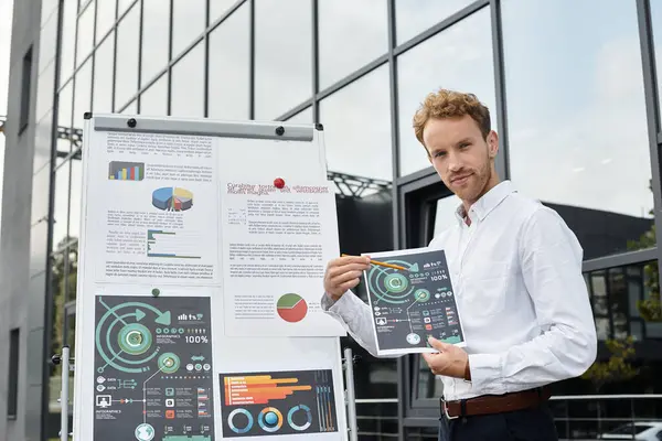 A redheaded businessman in a white shirt explains project data while standing in a contemporary office setting. — Stock Photo