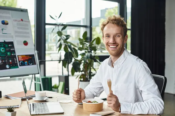 A redhead businessman in a white shirt smiles while enjoying lunch and focusing on his project in an office. — Stock Photo