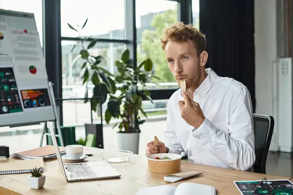 Un homme d'affaires roux concentré est engagé dans un projet tout en savourant son déjeuner dans un espace de bureau élégant. — Photo de stock