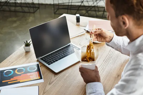 Un uomo d'affari in camicia bianca sta mangiando tagliatelle mentre si concentra sul suo progetto in un ufficio elegante. — Foto stock