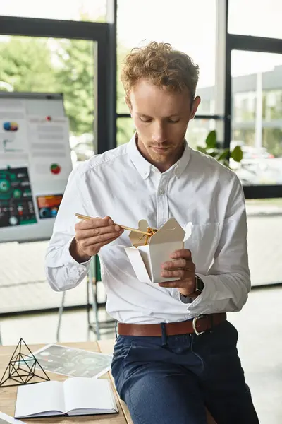 Un uomo d'affari professionista dai capelli rossi gode di cibo da asporto mentre lavora a un progetto in un ufficio elegante e moderno. — Foto stock