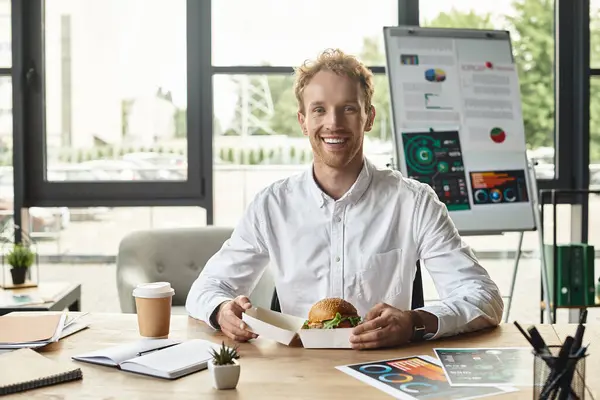 Une rousse dans une chemise blanche sourit tout en tenant un hamburger, concentré sur son projet dans un bureau élégant. — Photo de stock