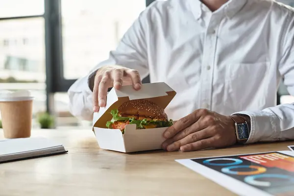 A man in a white shirt takes a moment to savor a burger while working in a contemporary office setting. — Stock Photo