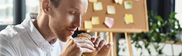 A focused businessman with red hair relishes a burger in his contemporary office while working on a project. — Stock Photo