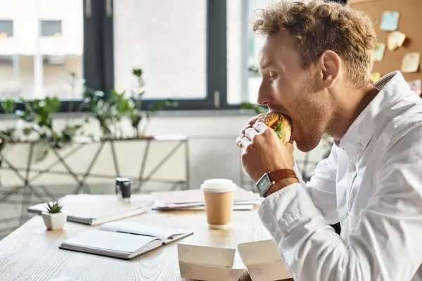The redhead entrepreneur takes a break to savor his burger in a contemporary workspace, balancing work and meal. — Stock Photo