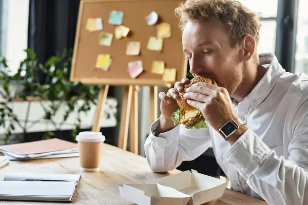 A redheaded businessman takes a break, relishing a delicious burger while working on a project. — Stock Photo