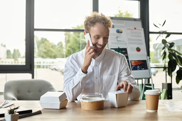 In a stylish office, a redhead man happily talks on the phone and enjoys his lunch during a project discussion. — Stock Photo