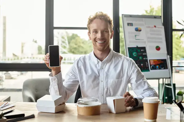 Une rousse en chemise blanche sourit en tenant un téléphone, concentrée sur un projet dans un cadre de bureau contemporain. — Photo de stock