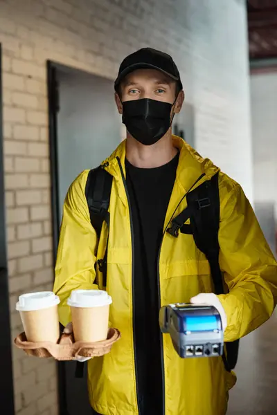 A delivery man in a black mask carries coffee cups into an office building, ensuring freshness and safety. — Stock Photo