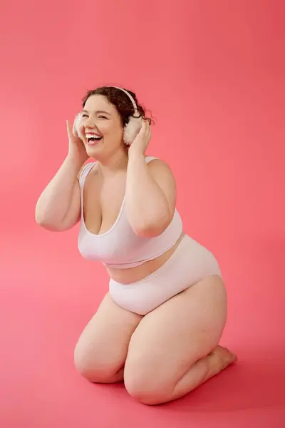 Eine fröhliche Plus-Size-Frau kniet auf einer weichen Oberfläche und genießt Musik mit kuscheligen Kopfhörern. — Stockfoto