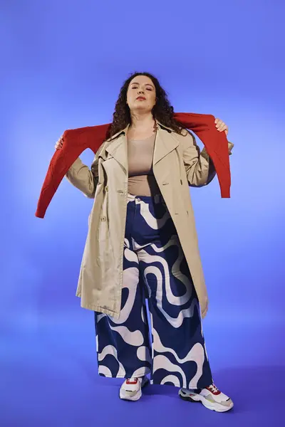 A confident plus size woman showcases a red sweater and stylish pants against a blue backdrop. — Stock Photo