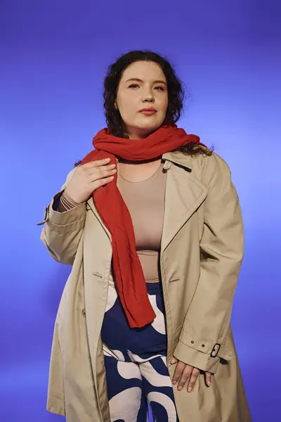 Una donna fiduciosa plus size in un trench e sciarpa rossa si erge contro il blu vibrante. — Foto stock
