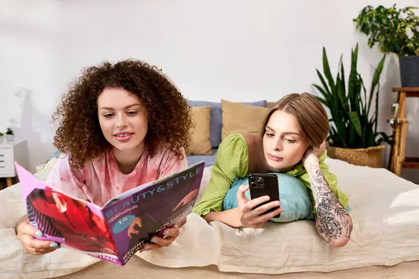 Duas mulheres felizes relaxar em uma cama acolhedora, desfrutando de uma revista e telefone em um espaço tranquilo. — Fotografia de Stock
