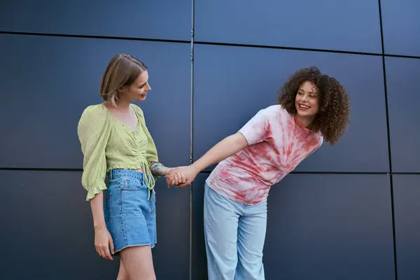 Two young women enjoy each others company, laughing and holding hands against a modern backdrop. — Stock Photo
