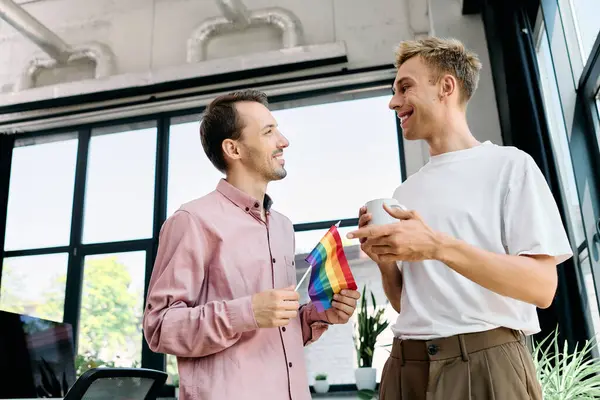 Two men smile and engage in conversation while holding a rainbow flag in a stylish office. — Stock Photo