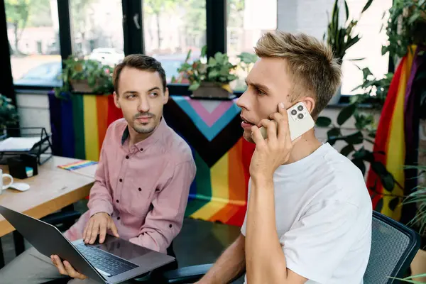 Two attractive men share a moment in a vibrant workspace, one on the phone while the other works. — Stock Photo