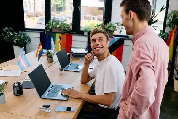 Deux hommes heureux s'engagent dans une discussion animée, entourés de drapeaux de fierté dans un bureau créatif. — Photo de stock