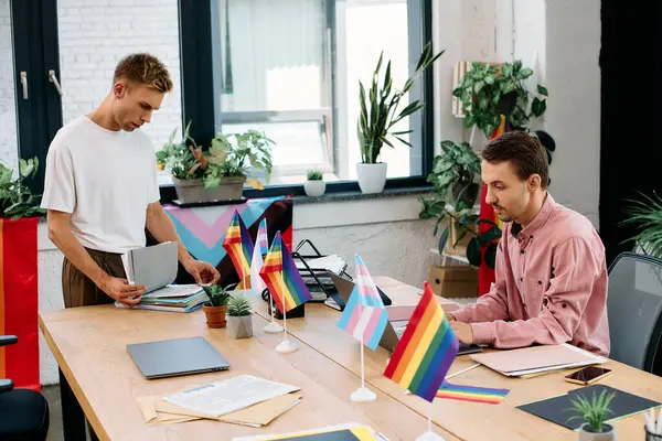 Two men sort paperwork in a lively, plant filled space adorned with pride flags. — Stock Photo