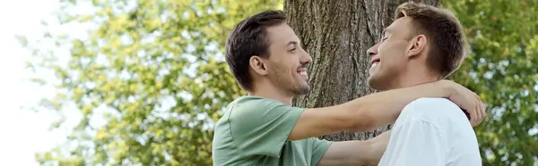 Two smiling men embrace joyfully in a vibrant park filled with greenery around them. — Stock Photo