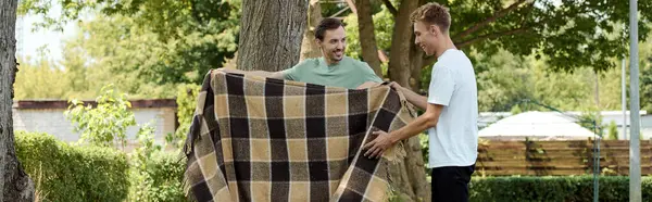Two smiling men share a light moment as they arrange a blanket in a beautiful outdoor setting. — Stock Photo