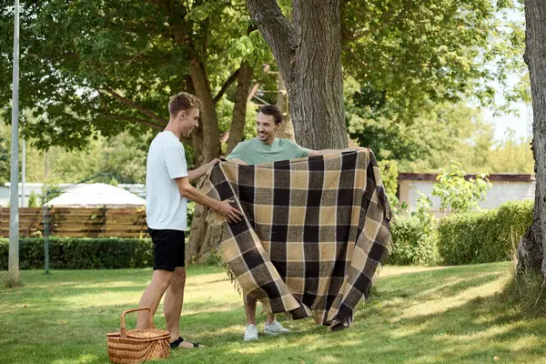 Two joyful men unfold a blanket together in a sunlit park, preparing for a delightful picnic. — Stock Photo