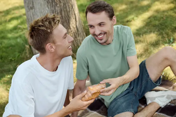 A happy couple sits on a blanket, sharing smiles and treats in a lush green park setting. — Stock Photo