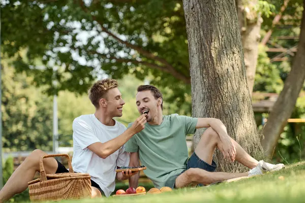 Two happy men enjoy a lovely picnic, feeding each other under a tree on a warm day. — Stock Photo
