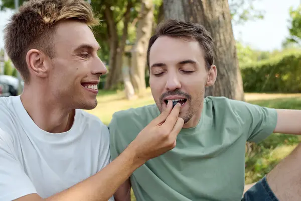Two smiling men enjoy a playful moment while feeding each other outdoors in a beautiful setting. — Stock Photo