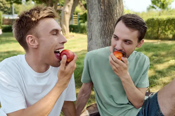 Dos hombres guapos comparten una tarde soleada, riendo y mordiendo melocotones jugosos. - foto de stock