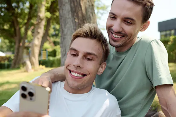 A cheerful gay couple smiles at their phone, sharing a delightful moment in the park. — Stock Photo