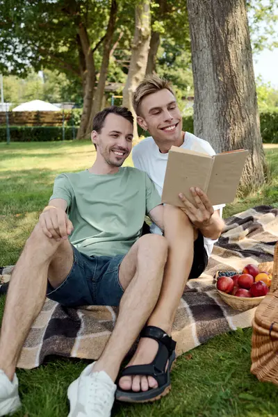 Two happy men share a picnic on a blanket, enjoying laughter and reading while outdoors. — Stock Photo