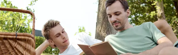 Two men sit closely in a sunny park, engrossed in a book, sharing smiles and laughter. — Stock Photo