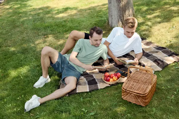 Two handsome men relax on a blanket, immersed in their books while savoring fruits under a tree. — Stock Photo
