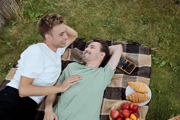 Two men relax on a checkered blanket, enjoying a sunny picnic with fruits and pastries. — Stock Photo