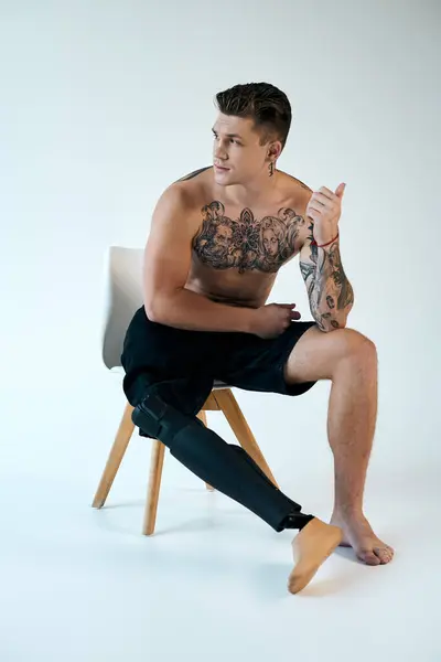 A young man with tattoos sits on a chair, confidently displaying his prosthetic leg in a sleek studio. — Stock Photo
