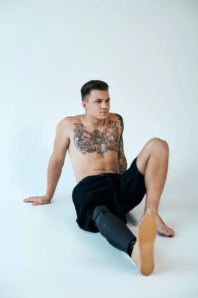 Um jovem tatuado senta-se no chão em um estúdio bem iluminado, mostrando sua perna protética e tatuagens.. — Fotografia de Stock