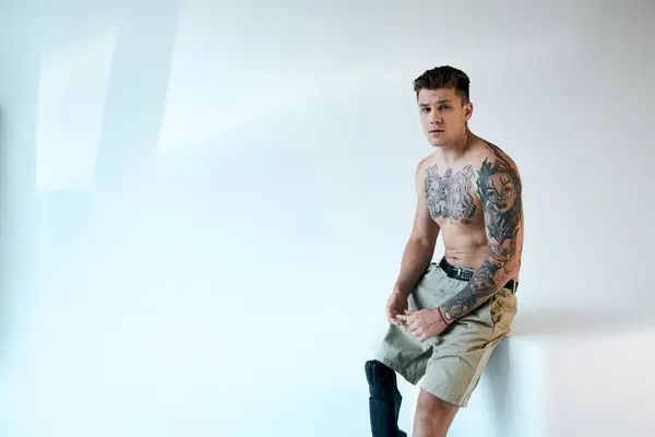 Un jeune homme confiant montre ses tatouages tout en s'appuyant contre un mur de studio propre, avec une jambe prothétique visible. — Photo de stock