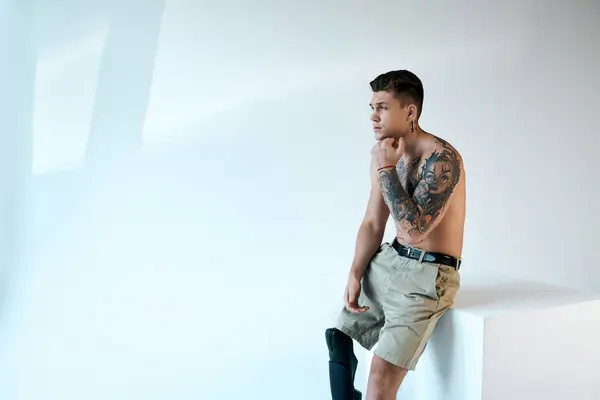 Um jovem tatuado com uma perna protética senta-se contemplativamente em um estúdio brilhante e minimalista. — Fotografia de Stock