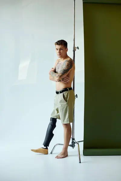 A young man with tattoos stands in a studio, showcasing his prosthetic leg while radiating confidence. — Stock Photo