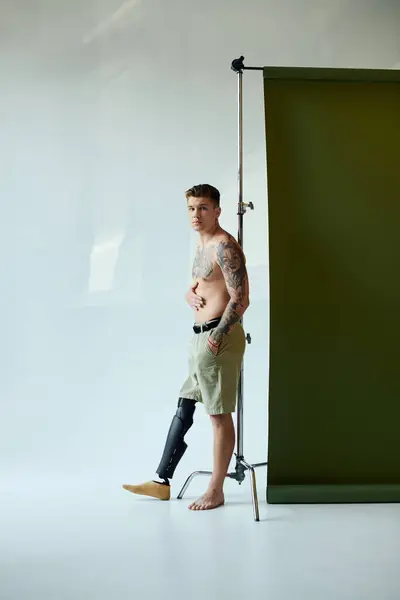 A young man with tattoos stands proudly in a studio, showcasing his prosthetic leg and relaxed style. — Stock Photo