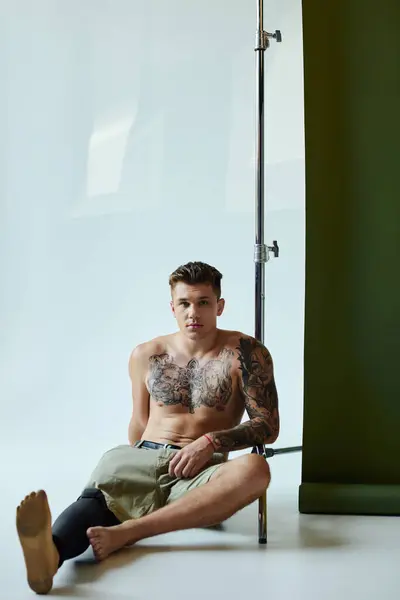 Ein tätowierter junger Mann sitzt vor einem Hintergrund und zeigt seine Beinprothese und seinen künstlerischen Ausdruck. — Stockfoto