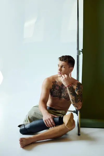 A young man with intricate tattoos sits thoughtfully in a studio, confidently highlighting his prosthetic leg. — Stock Photo