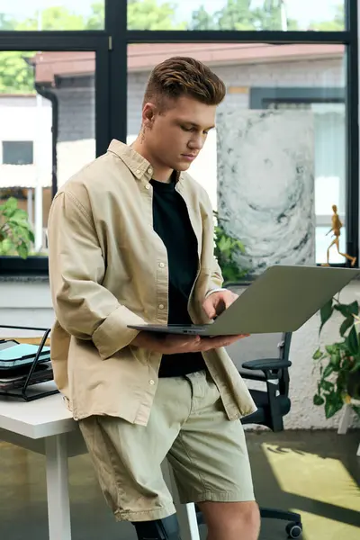 A young man with a prosthetic leg focuses intently on his laptop while standing in a sleek office space. — Stock Photo