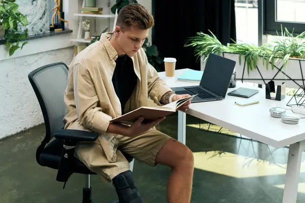 A young man with a prosthetic leg engrossed in reading a book at his contemporary office workspace. — Stock Photo
