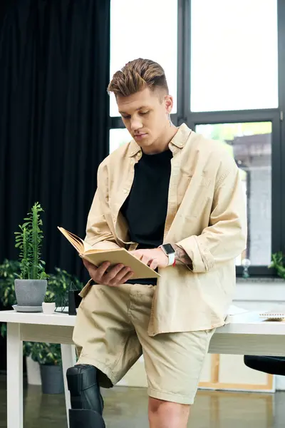 A motivated young man with a prosthetic leg is engrossed in reading a book in a stylish office setting. — Stock Photo