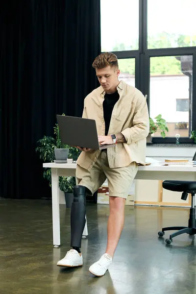 A determined young man using a laptop stands confidently in a contemporary office setting. — Stock Photo
